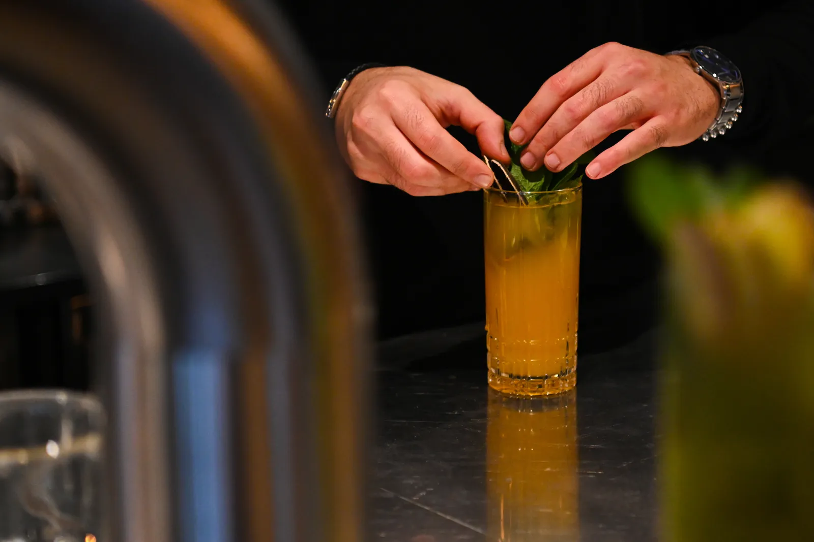 A bartender's hands setting mint into a tall amber cocktail on the bar counter