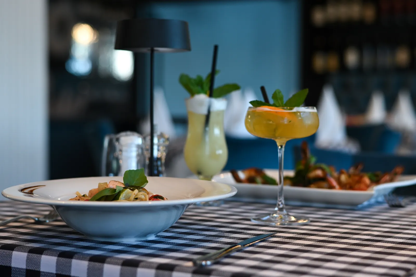 A bowl of pasta and two cocktails on a checked tablecloth, the dark bar behind