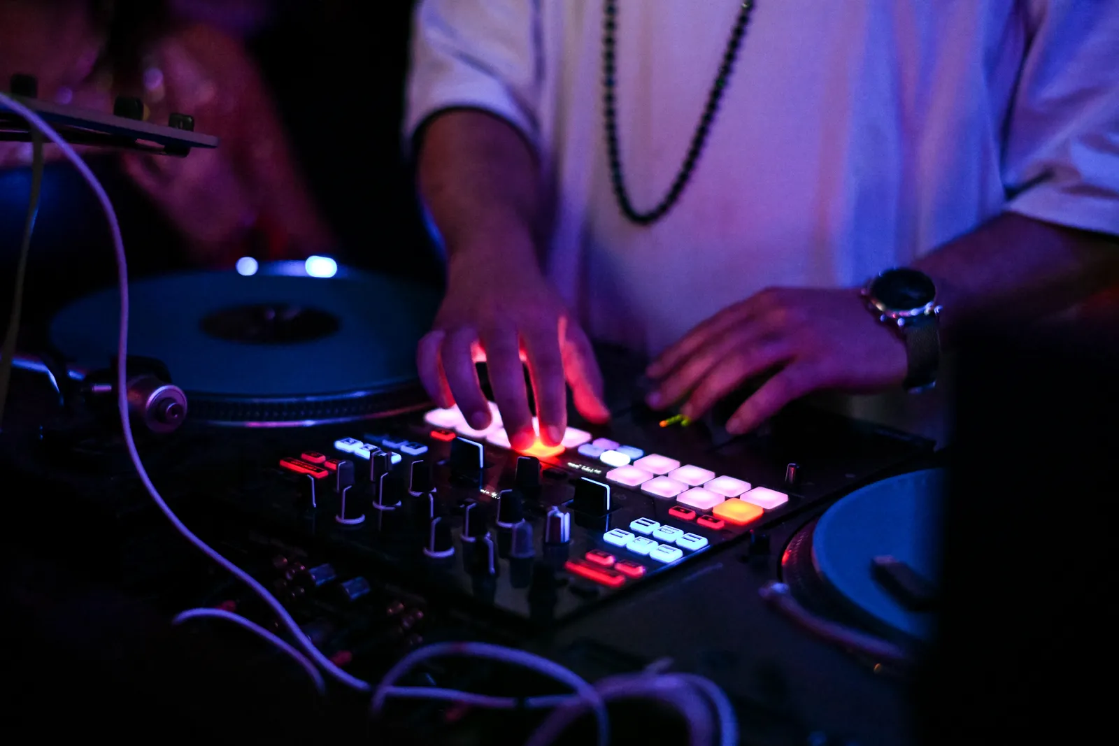 Hands over a DJ mixer and turntable, the pads lit red and the room in deep blue behind