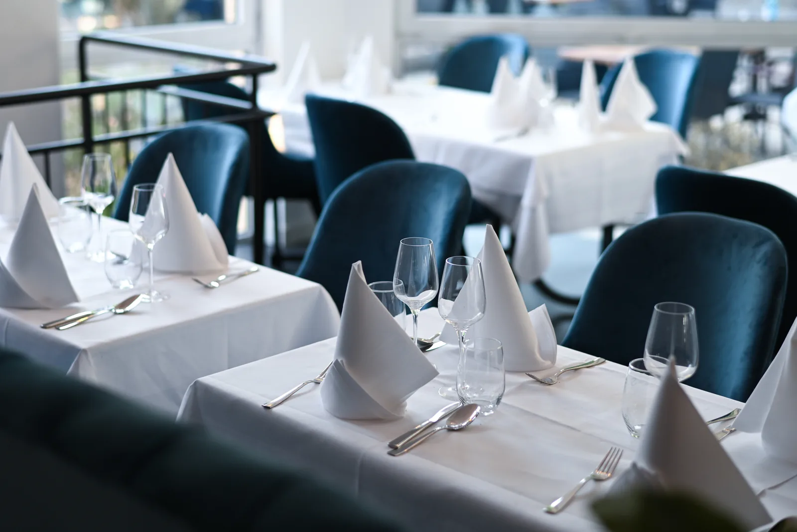 A dining room of tables laid with white linen and folded napkins, deep-blue velvet banquettes along the windows