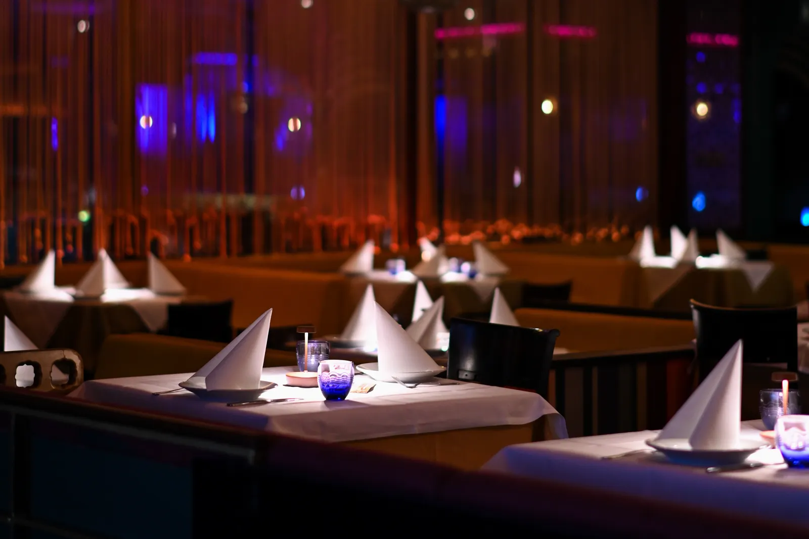 The dining room after dark: tables laid with white linen and folded napkins, tealights burning, the room lit blue and amber behind