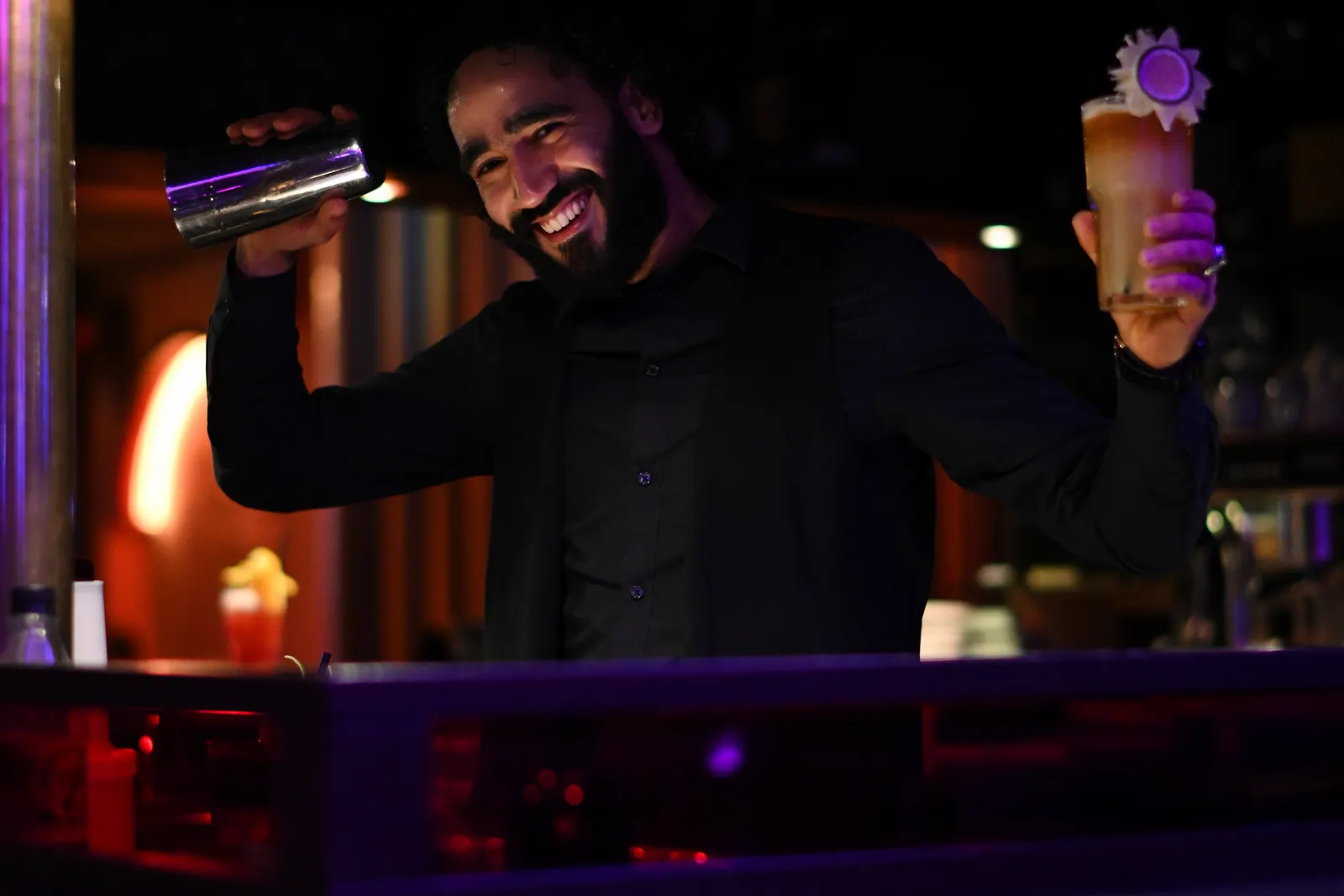 A bartender shaking a tin in one hand and holding a finished cocktail in the other, laughing behind the bar